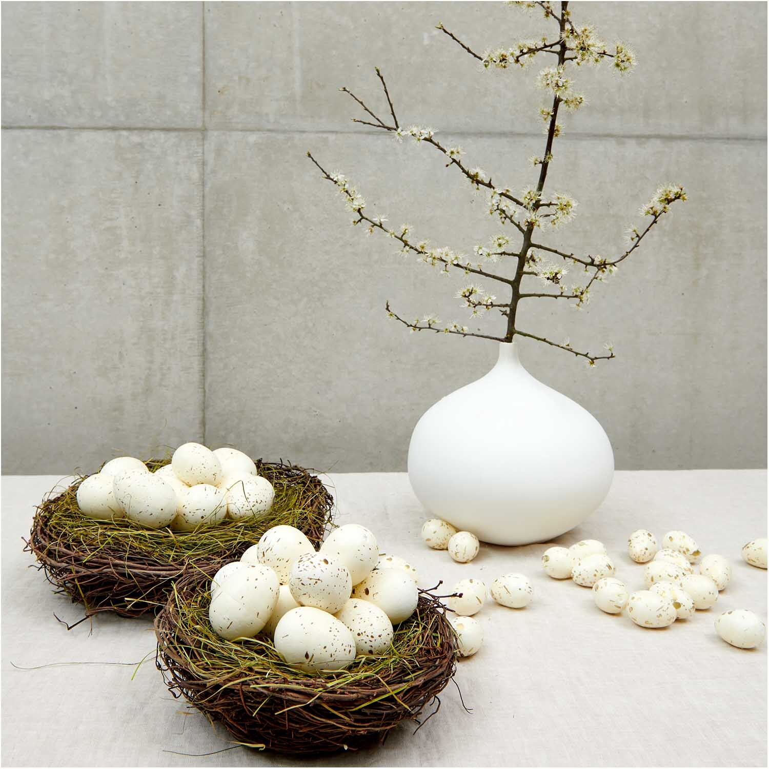 Eier im Netz 6cm 9 Stück von Rico Design Eier im Netz 6cm 9 Stück von Rico Design: Dekorative weiße Eier in Nestern neben einer Vase mit blühenden Zweigen auf einem Tisch.