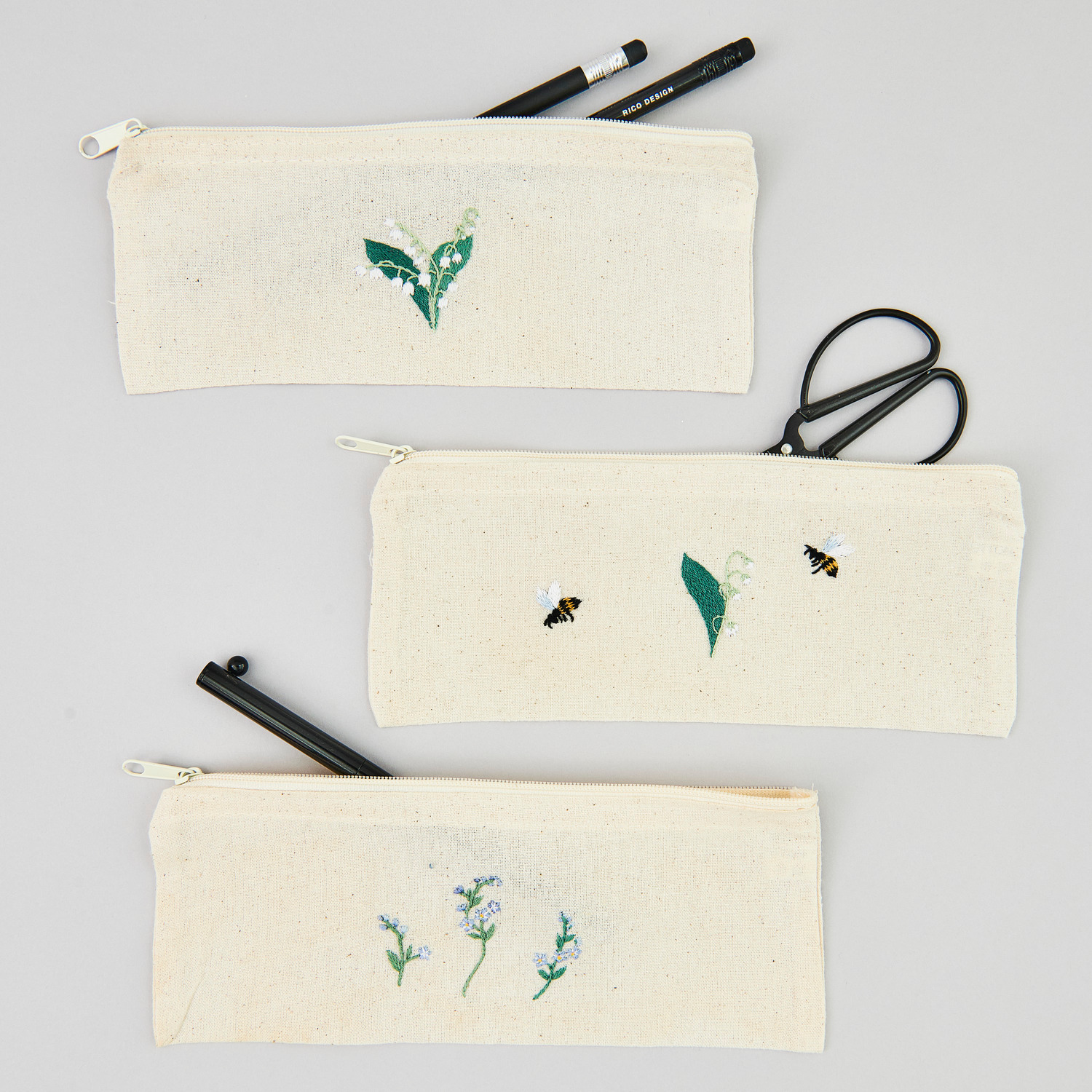 Stick and Stitch Blümchen: Drei naturfarbene Etuis mit floralen Motiven, gefüllt mit Stiften und einer Schere.