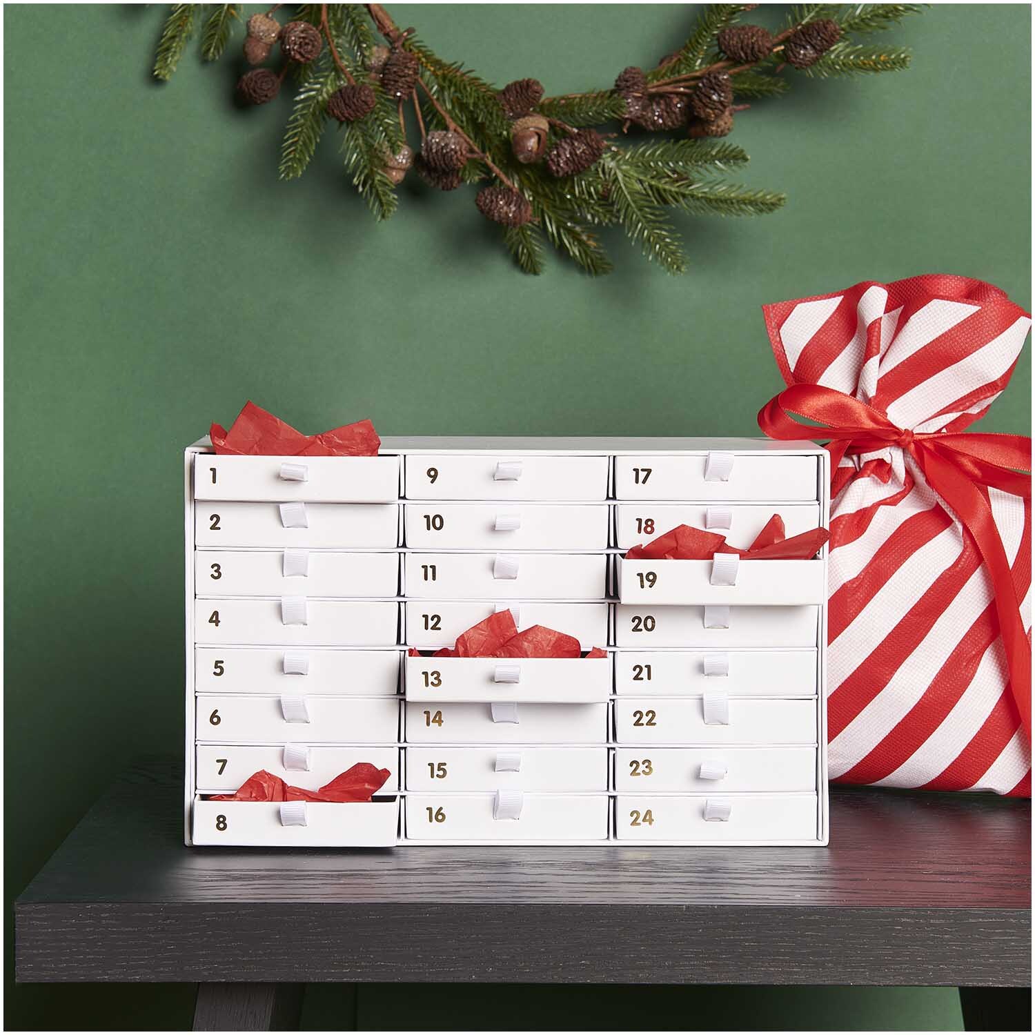 Adventskalender Box weiß-gold 16x26x9,5cm von Rico Design Adventskalender Box weiß-gold von Rico Design auf einem Tisch, dekoriert mit Tannenzweigen und einem gestreiften Geschenkbeutel.