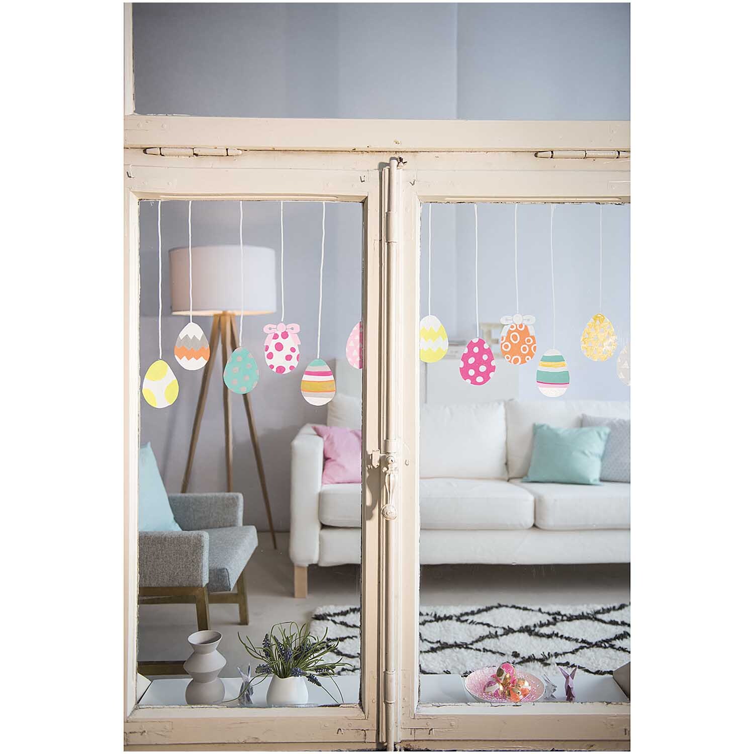 Vorlagenmappe Ostern Fenster- und Glasmalerei mit Kreidestiften von Rico Design Vorlagenmappe Ostern Fenster- und Glasmalerei mit bunten Eiern, dekoriert mit Kreidestiften, im Wohnzimmer mit Sofa und Kissen.