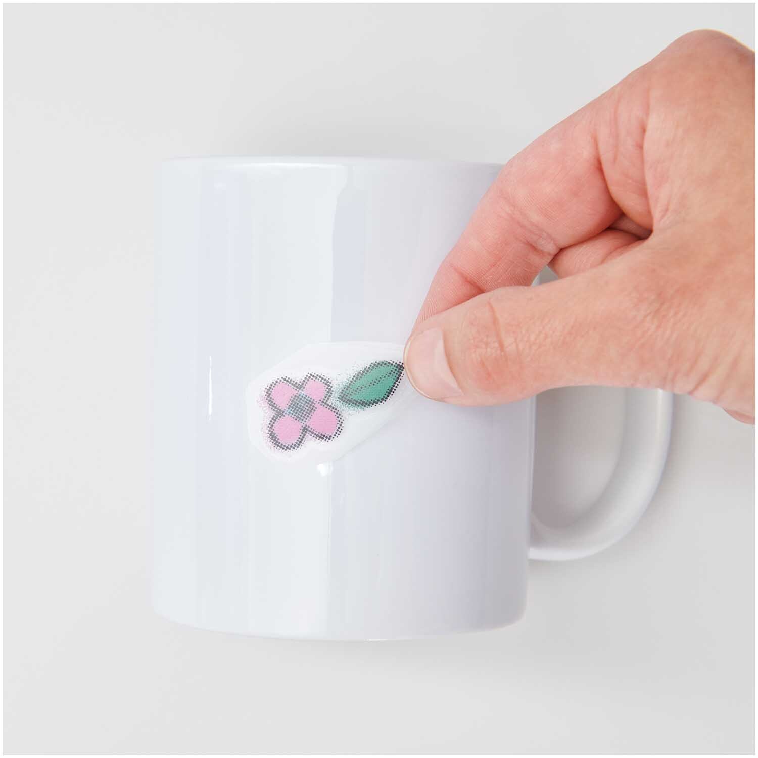 Transfermotiv Spray-Mix 10 Stück von Rico Design Transfermotiv Spray-Mix 10 Stück: Eine Hand klebt ein florales Motiv auf eine weiße Tasse. Marke: Rico Design.