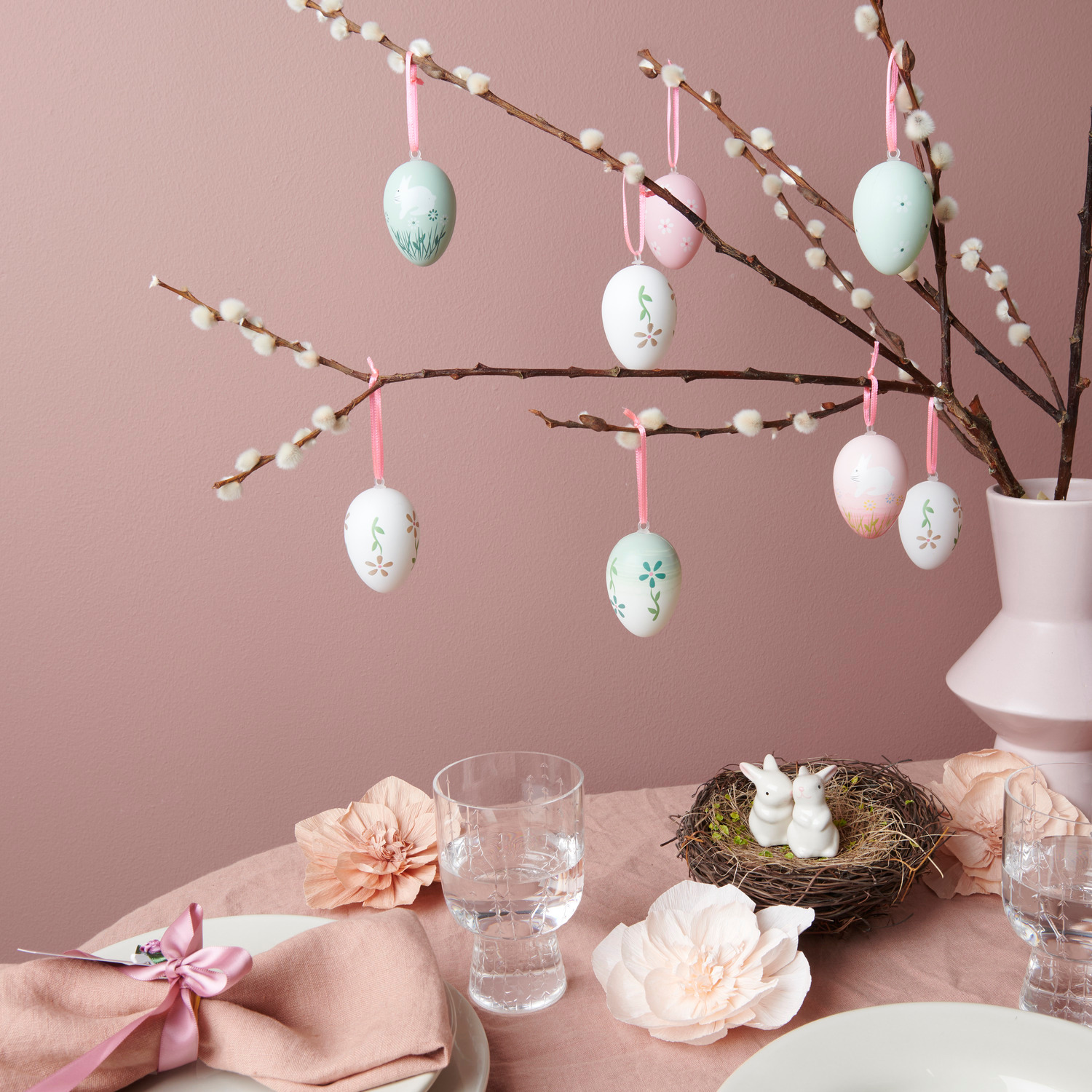 Deko-Eier Grün und Rosa hängen an Zweigen in einer Vase. Tisch mit Keramik Hasen und Krepppapier Blüten dekoriert.