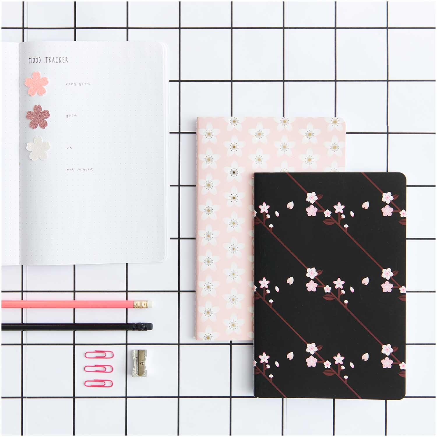 Paper Poetry Notizbücher mit Kirschblütenmuster in rosa und schwarz, umgeben von Stiften und Büroklammern auf kariertem Hintergrund.