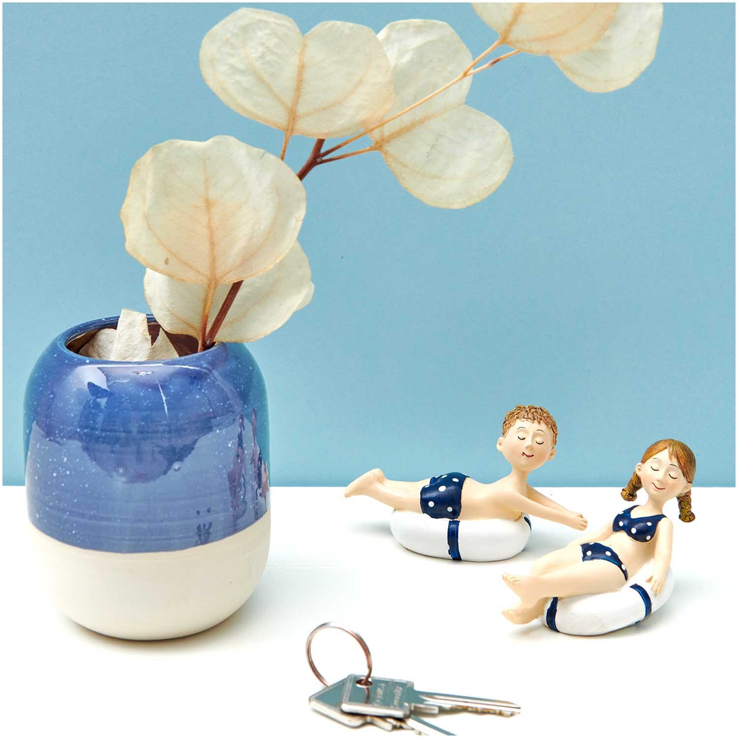 Figurine déco fillette avec bouée 9x6cm from Rico Design Deko-Figur Mädchen mit Schwimmreifen 9x6cm von Rico Design neben blauem Keramiktopf mit Pflanze.