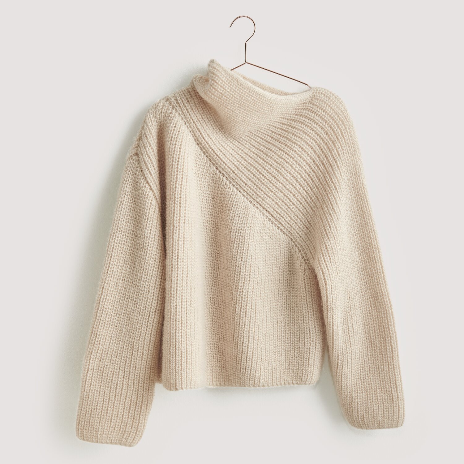 Luxury Merino Alpaca Dream aran 50g 125m Farbe Creme von Rico Design Strickset Pullover Modell 01 aus Luxury Knits: Beigefarbener Pullover mit asymmetrischem Strickmuster, hängend an einem Kleiderbügel.