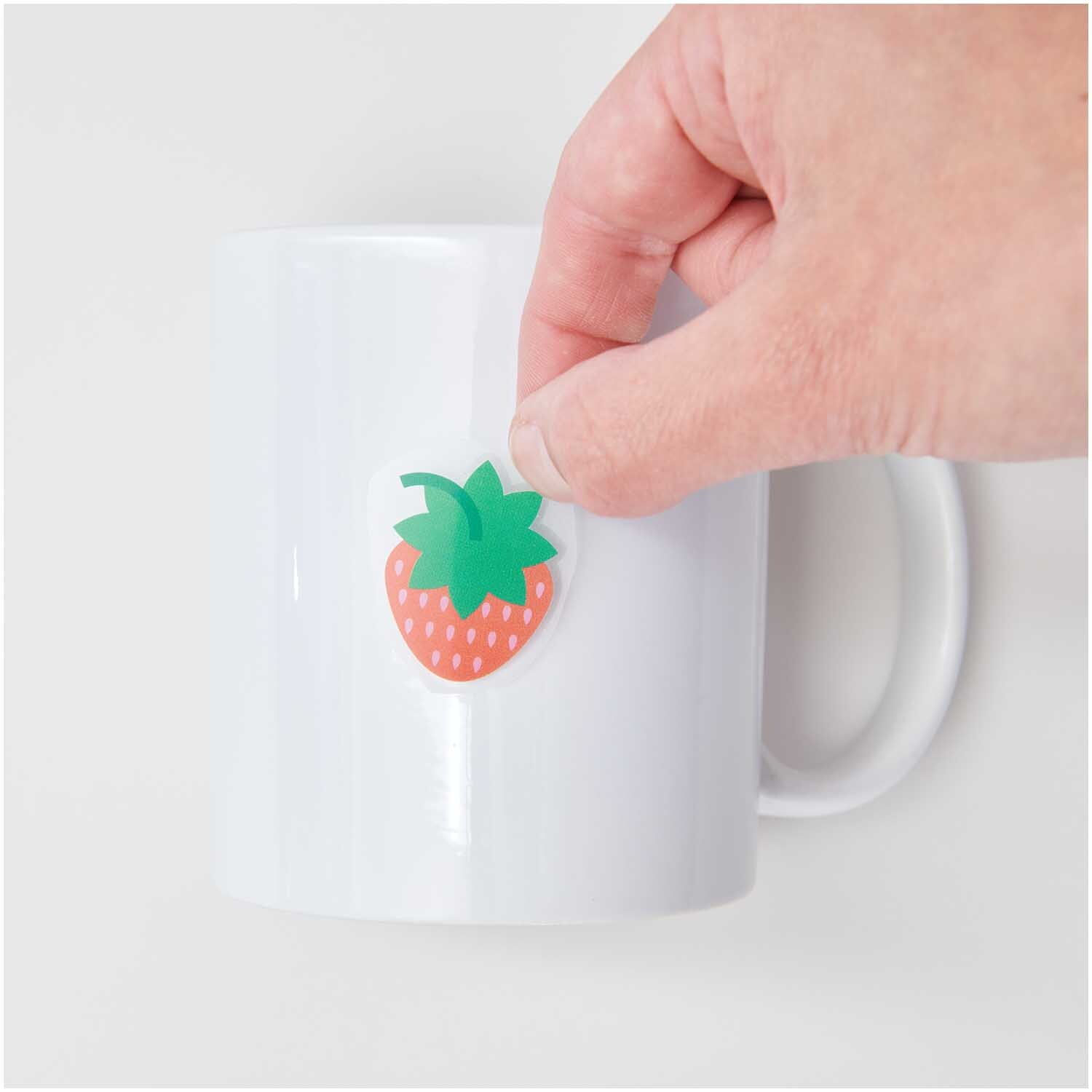Transfermotiv Früchte & Blumen: Hand klebt Erdbeer-Motiv auf weiße Tasse. Set von 15 Stück von Rico Design.