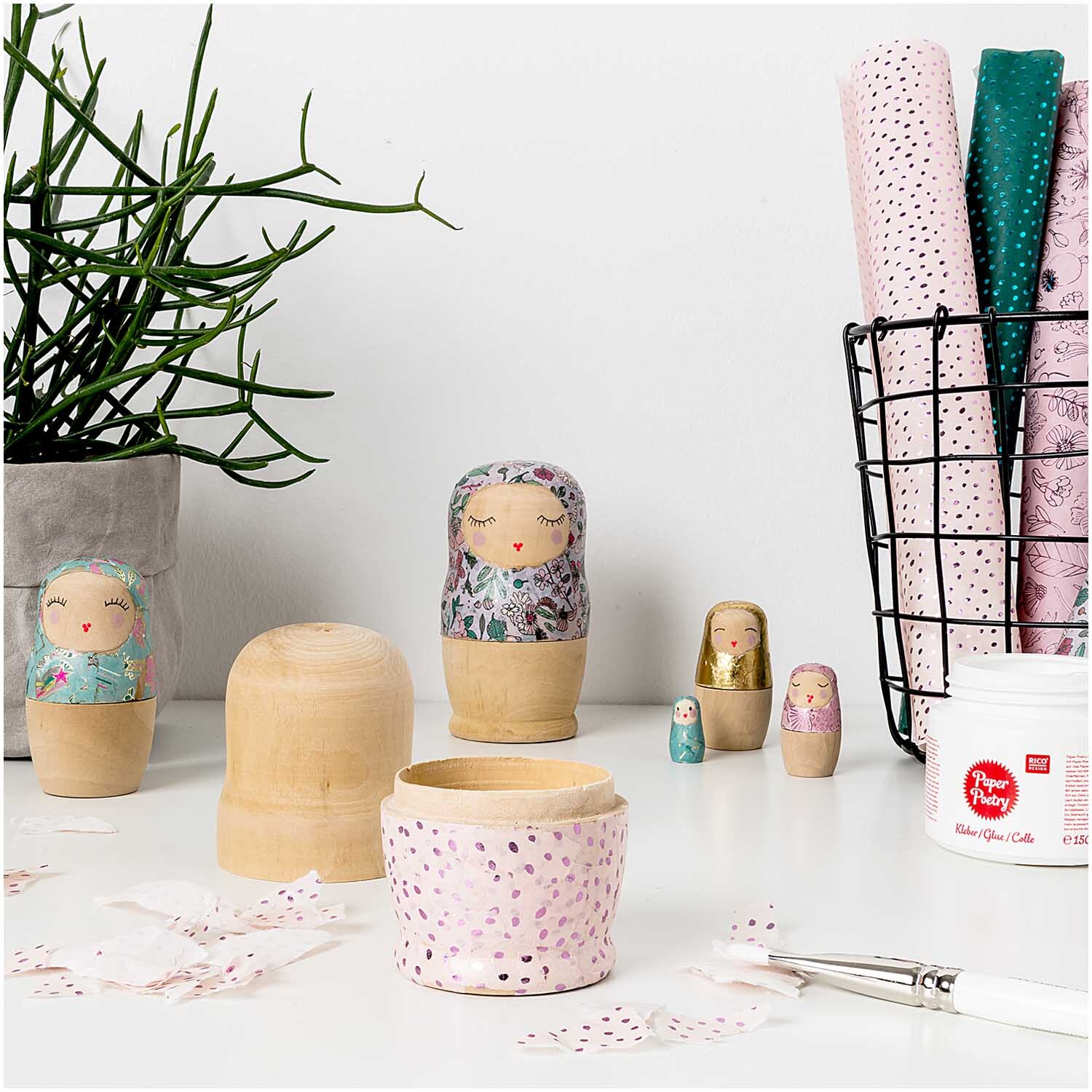 Paper Poetry Kleber von Rico Design neben bemalten Matroschka-Puppen und Bastelmaterialien.