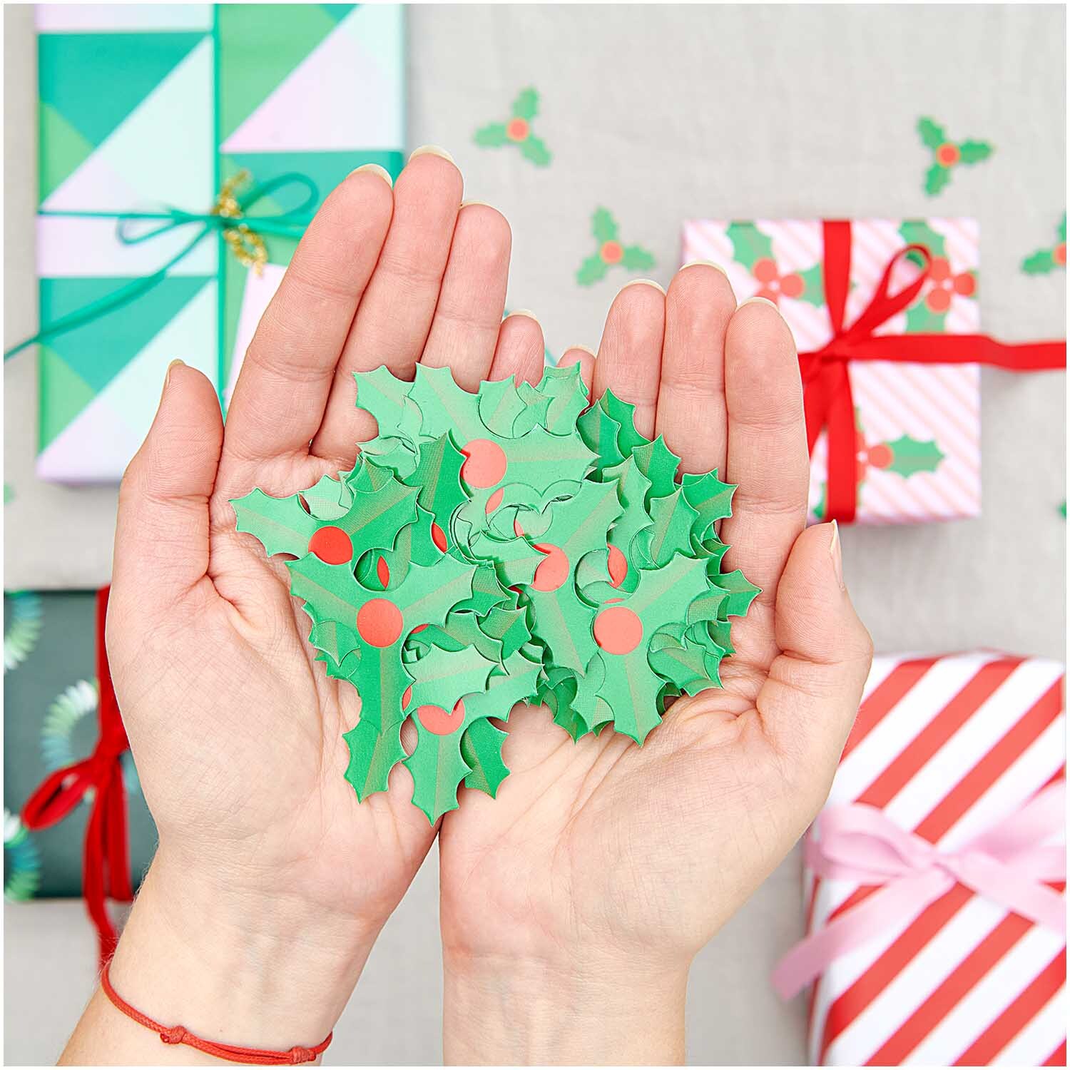 Streudeko Ilex 40g von Rico Design Streudeko Ilex 40g: Hände halten grüne Ilex-Blätter mit roten Beeren, umgeben von Geschenken.