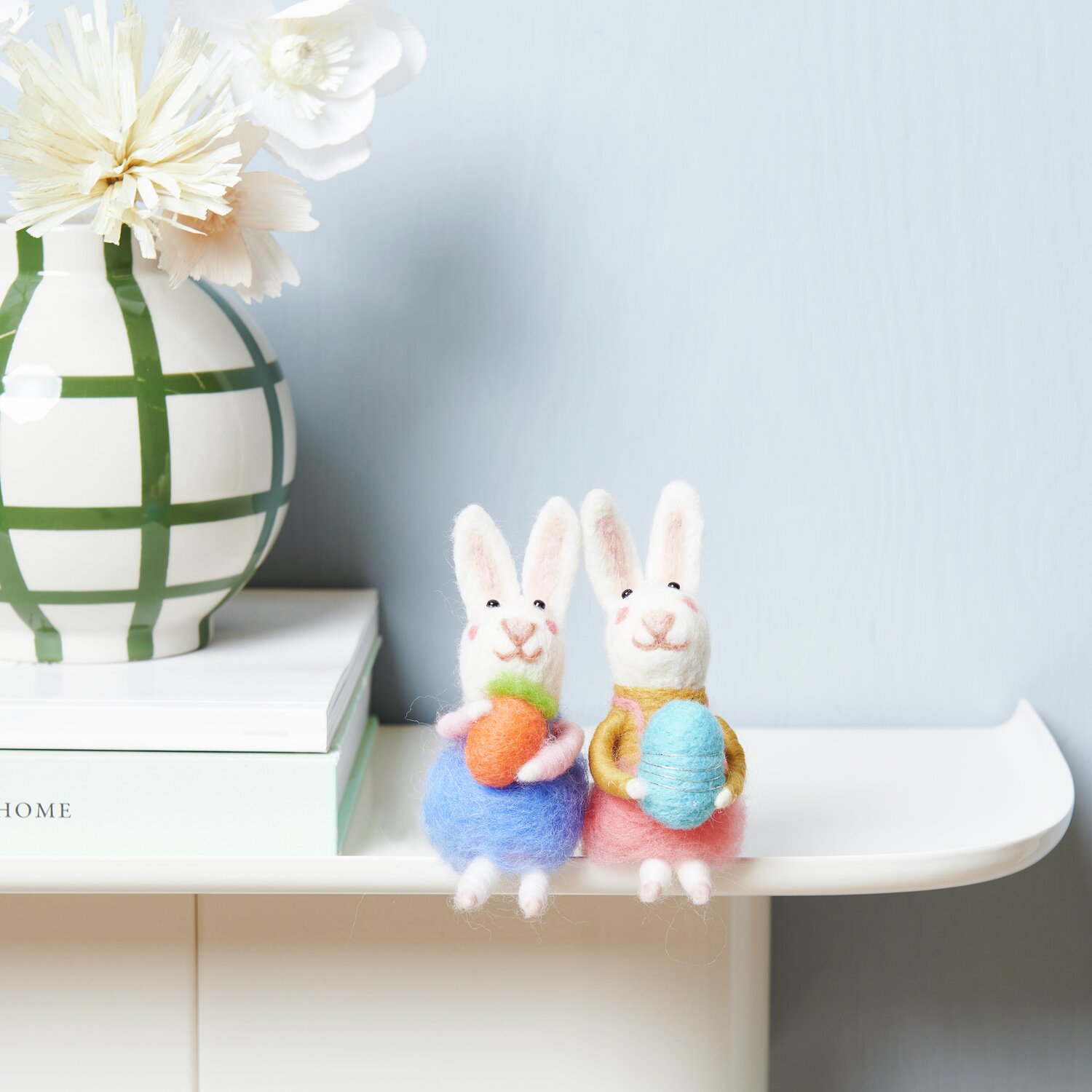 Aufhänger Filz-Hase mit Möhre 14x5x6,5cm von Rico Design Aufhänger Filz-Hase mit Osterei: Zwei Filz-Hasen mit bunten Eiern sitzen auf einem Regal, dekorativ neben einer Vase.