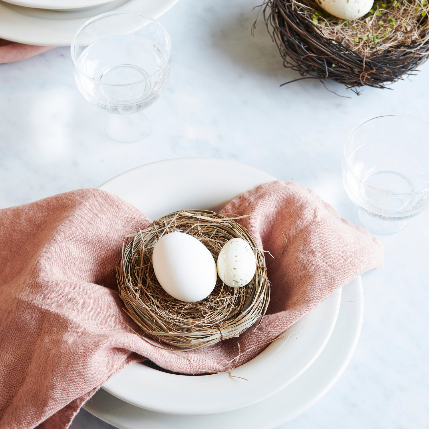 Eier creme-gold 6cm 6 Stück von Rico Design Naturnest mit Gras und creme-goldenen Eiern auf einem gedeckten Tisch, umgeben von Geschirr und Gläsern.