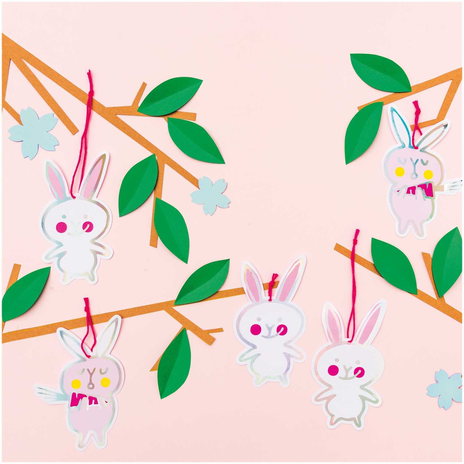 Paper Poetry Papieranhänger Bunnies 8 Stück: Weiße Häschen-Anhänger von Rico Design an Zweigen mit grünen Blättern und rosa Hintergrund.
