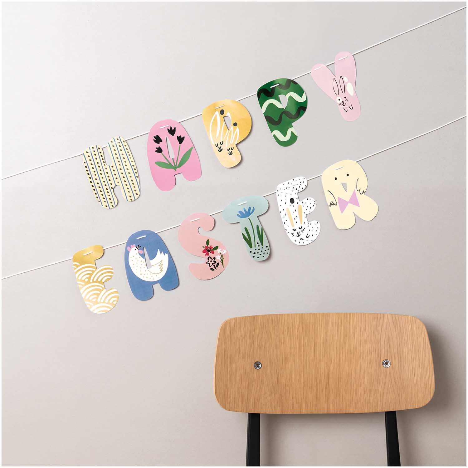 Paper Poetry Girlande Happy Easter 3m von Rico Design Paper Poetry Girlande Happy Easter 3m: Bunte Buchstaben-Girlande mit Osterdesigns, dekorativ an der Wand über einem Holzstuhl aufgehängt.