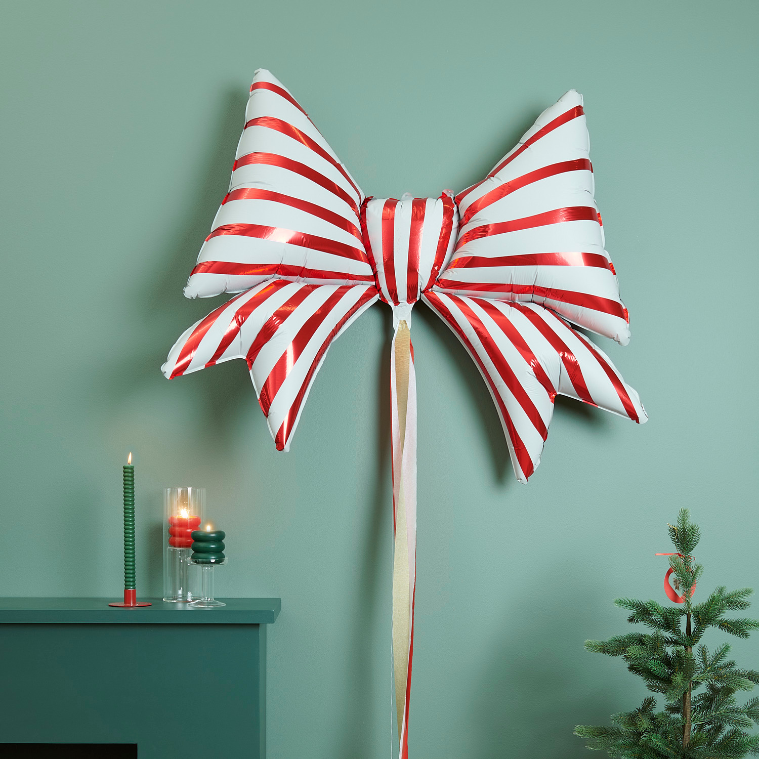 Folienballon Schleife Rot-Weiß 100x80x22cm 1 Stück von Rico Design Folienballon Schleife Rot-Weiß von Rico Design, dekorativ an einer grünen Wand mit Kerzen und Tannenbaum.