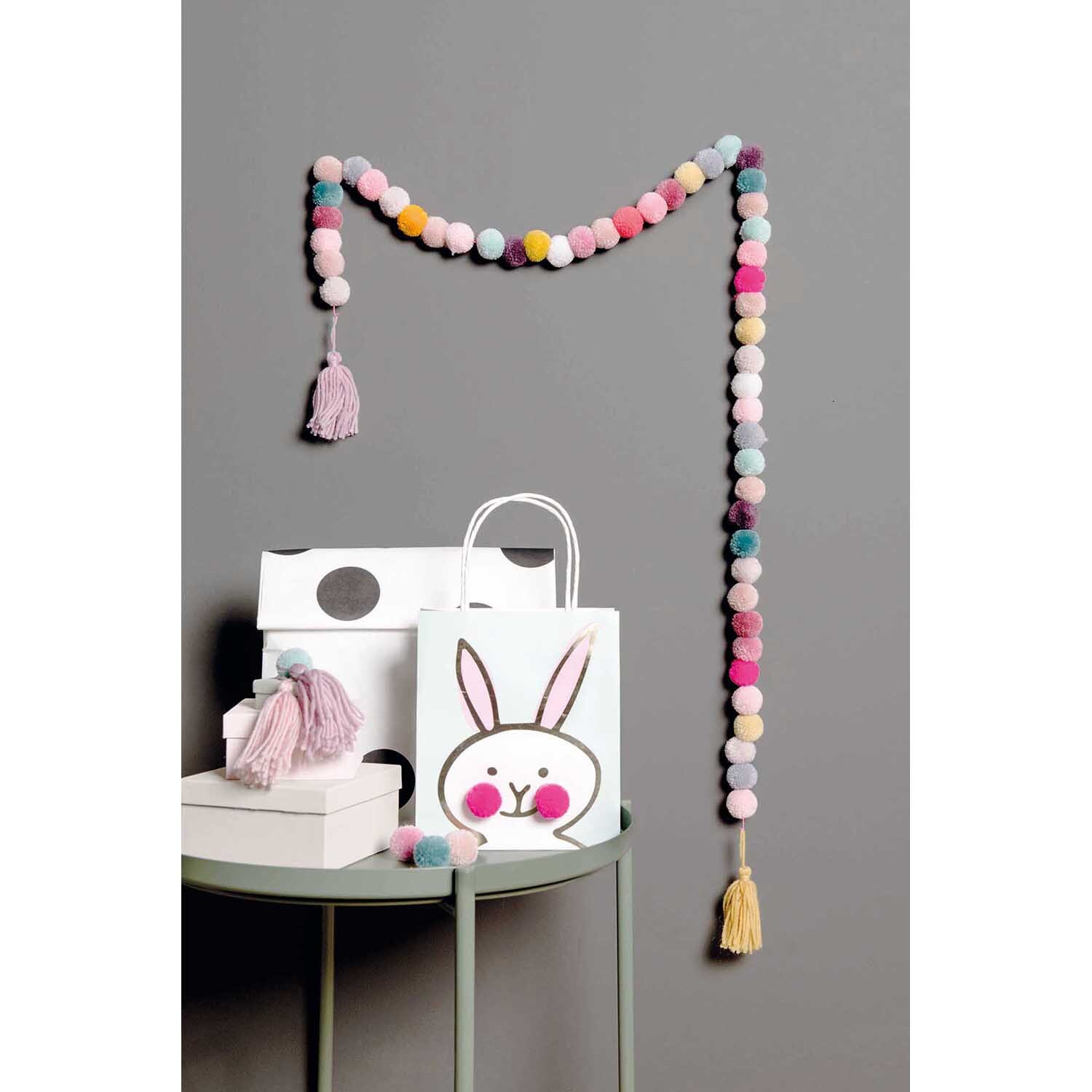 Garn Pompons 24 Stück Farbe Natur Mix von Rico Design Garn Pompons 24 Stück: Bunte Pompon-Girlande von Rico Design, dekorativ an grauer Wand hängend.