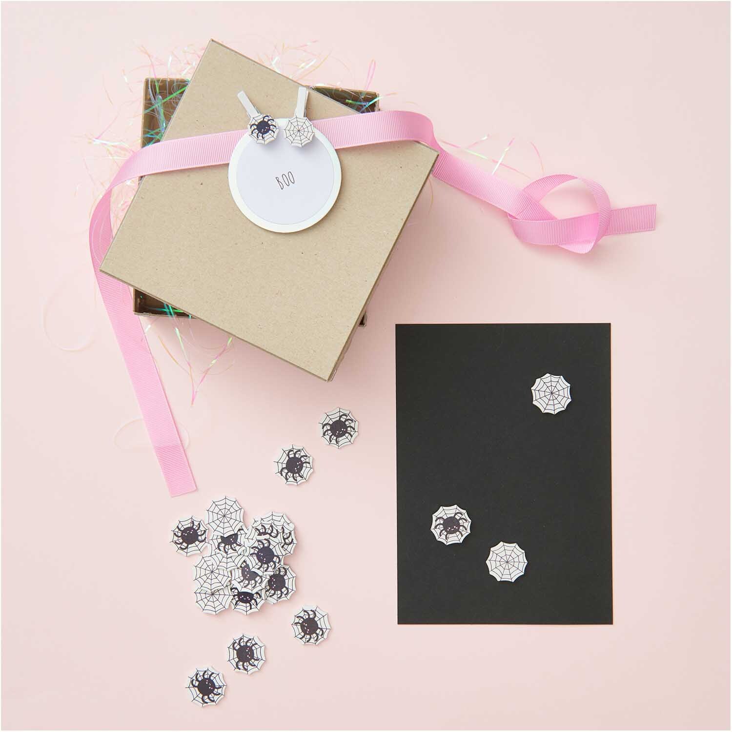 Holzsticker Spinnennetz Mix schwarz-weiß 12 Stück von Rico Design Holzsticker Spinnennetz Mix schwarz-weiß 12 Stück auf schwarzem Karton, dekoriert mit Geschenkbox und rosa Band. Ideal für kreative Bastelprojekte.