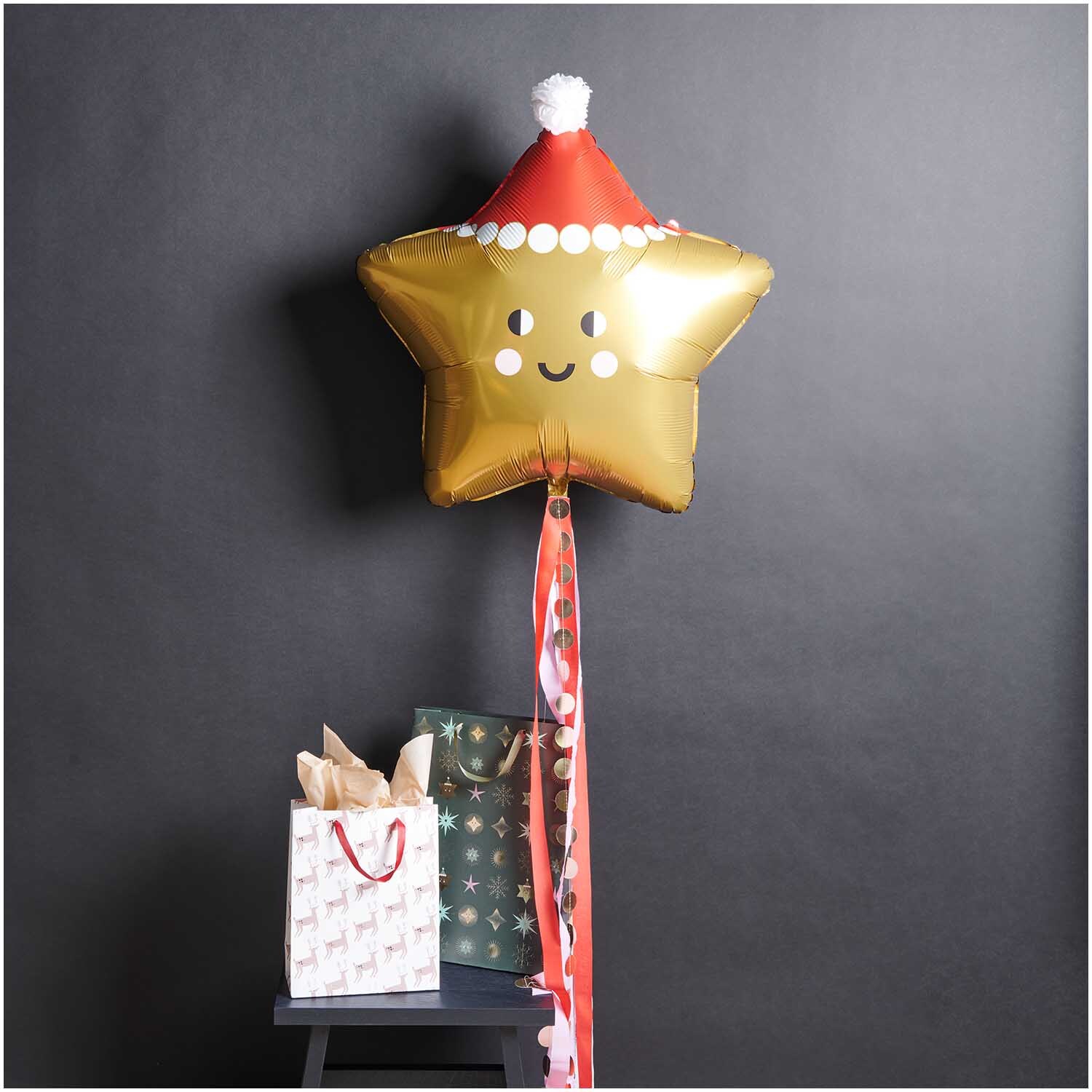 Folienballon Weihnachten Stern ca. 85x88,5cm von Rico Design Folienballon Weihnachten Stern in Gold mit Weihnachtsmütze, dekoriert neben Geschenktüten.
